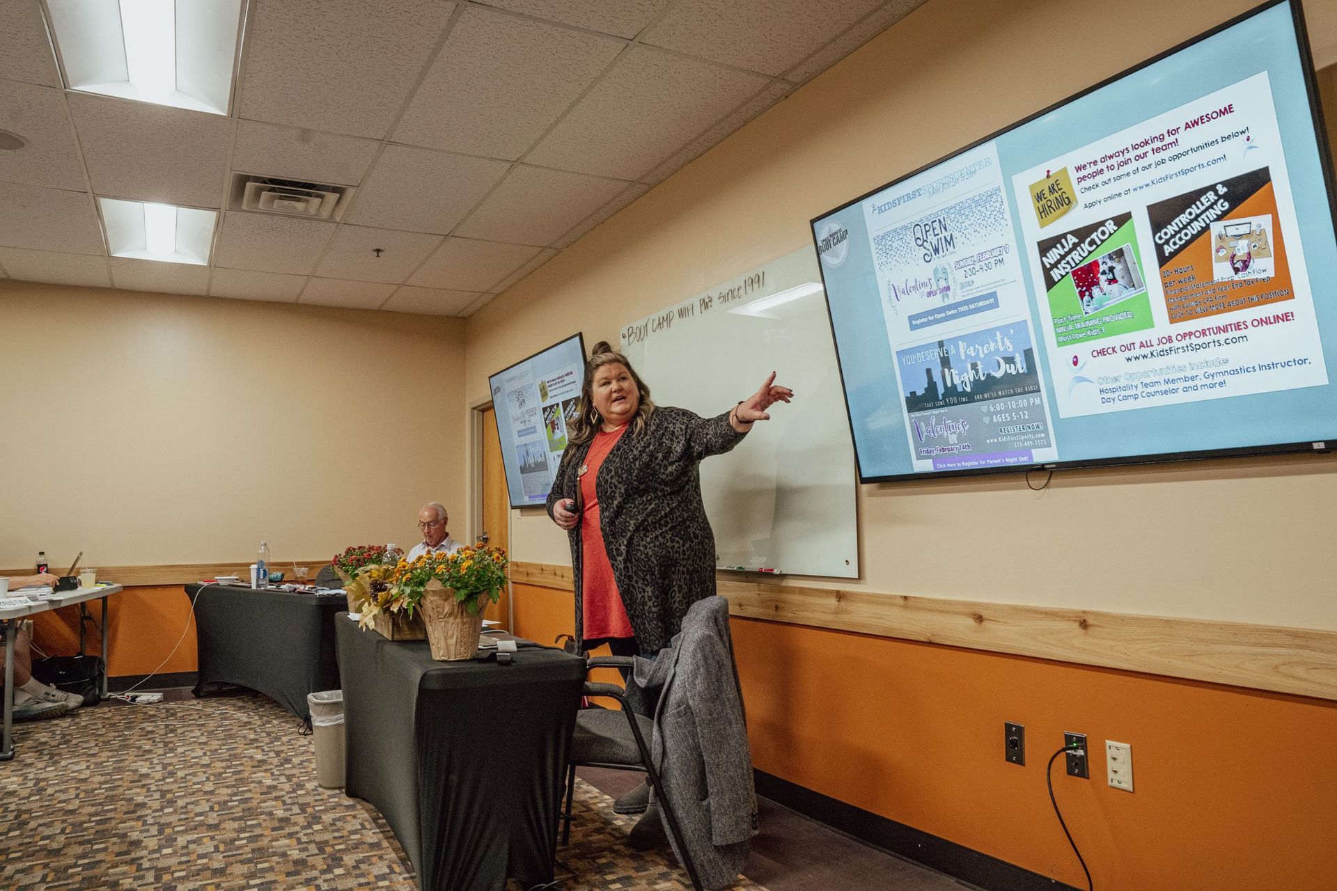 Presenter standing beside screen in a classroom, gesturing toward slides during a presentation.