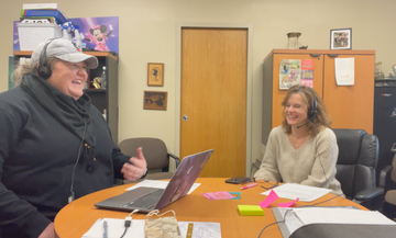 Two people talking in an office, with a laptop on the table and papers spread out.