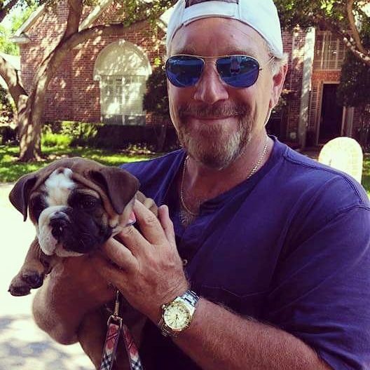 Man in blue shirt and sunglasses holding a bulldog puppy outdoors.