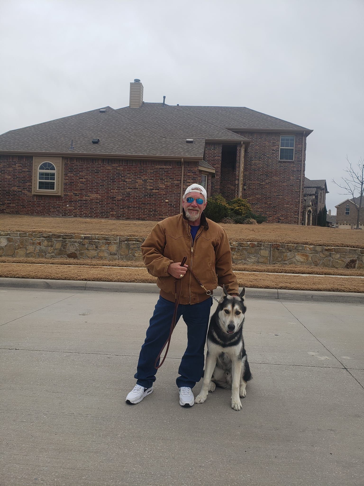 Man in brown jacket and husky dog posing in front of a brick house on a street.