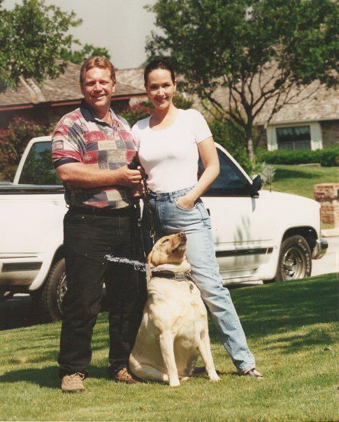 Man and woman standing with yellow Labrador, in front of a white truck, on a lawn.