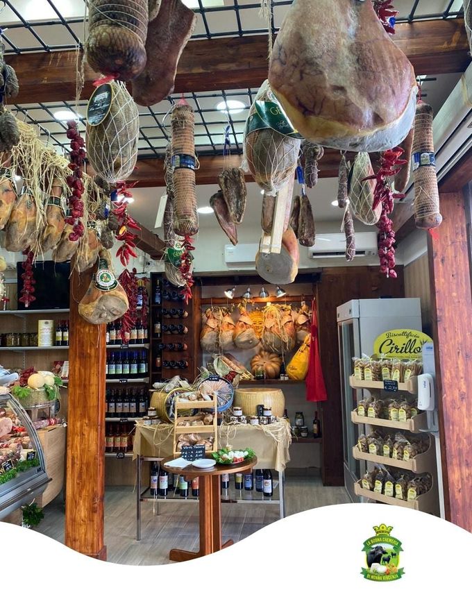 Interno di una gastronomia, con salumi esposti appesi al soffitto, banconi con cibo e scaffali con prodotti.