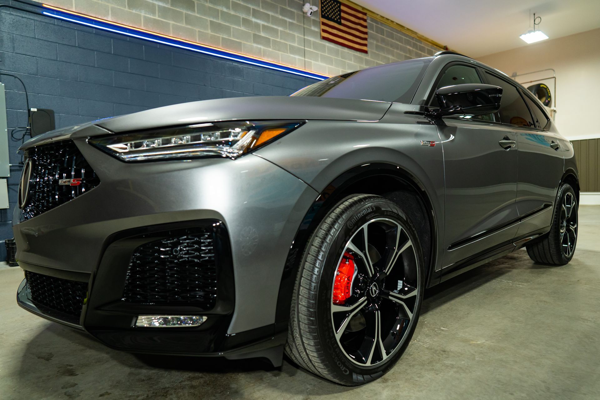 A gray Acura MDX Type S parked in an indoor garage with an American flag displayed on the block wall.
