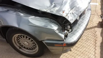 A damaged silver car showing a dented front fender, crumpled hood, and missing headlight on the passenger side.