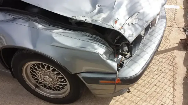 A damaged silver car showing a dented front fender, crumpled hood, and missing headlight on the passenger side.