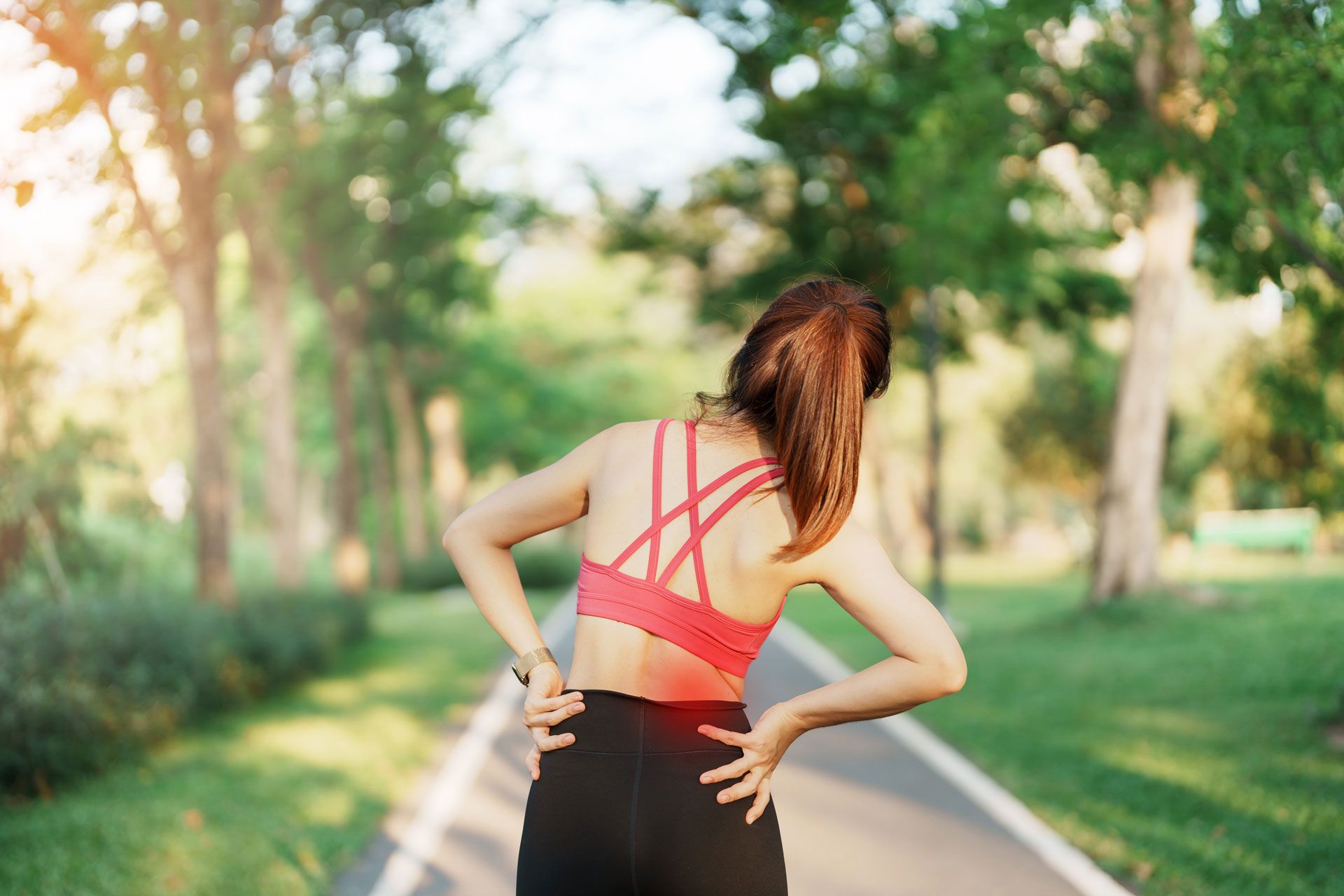 Woman in sportswear, holding her lower back in pain while standing on a path in a park.