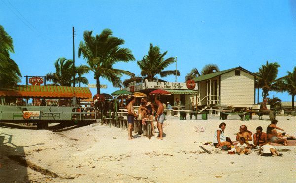 A group of people standing on a beach in front of a building that says ' coca cola ' on it