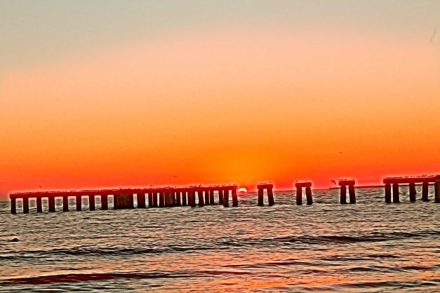 The sun is setting over the ocean with a pier in the foreground.
