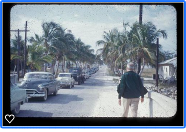 A man is walking down a street with cars parked on both sides