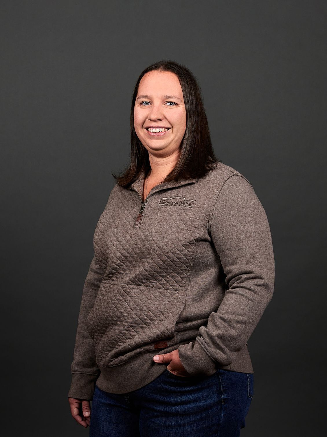 Woman in brown quilted sweater and jeans smiles, hand in pocket, against a gray background.