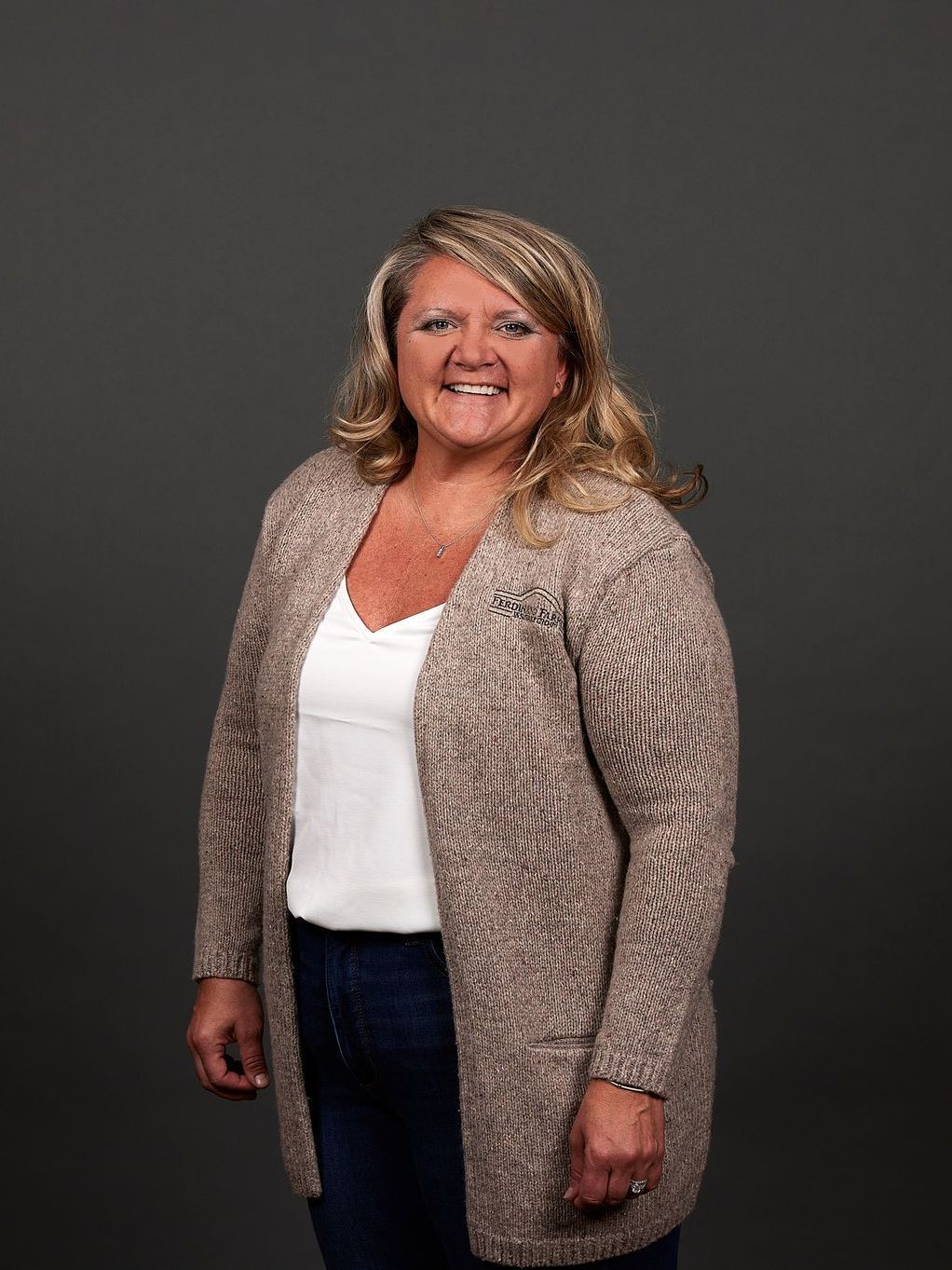 Woman in a cream sweater and white top, smiling, standing in front of a gray backdrop.
