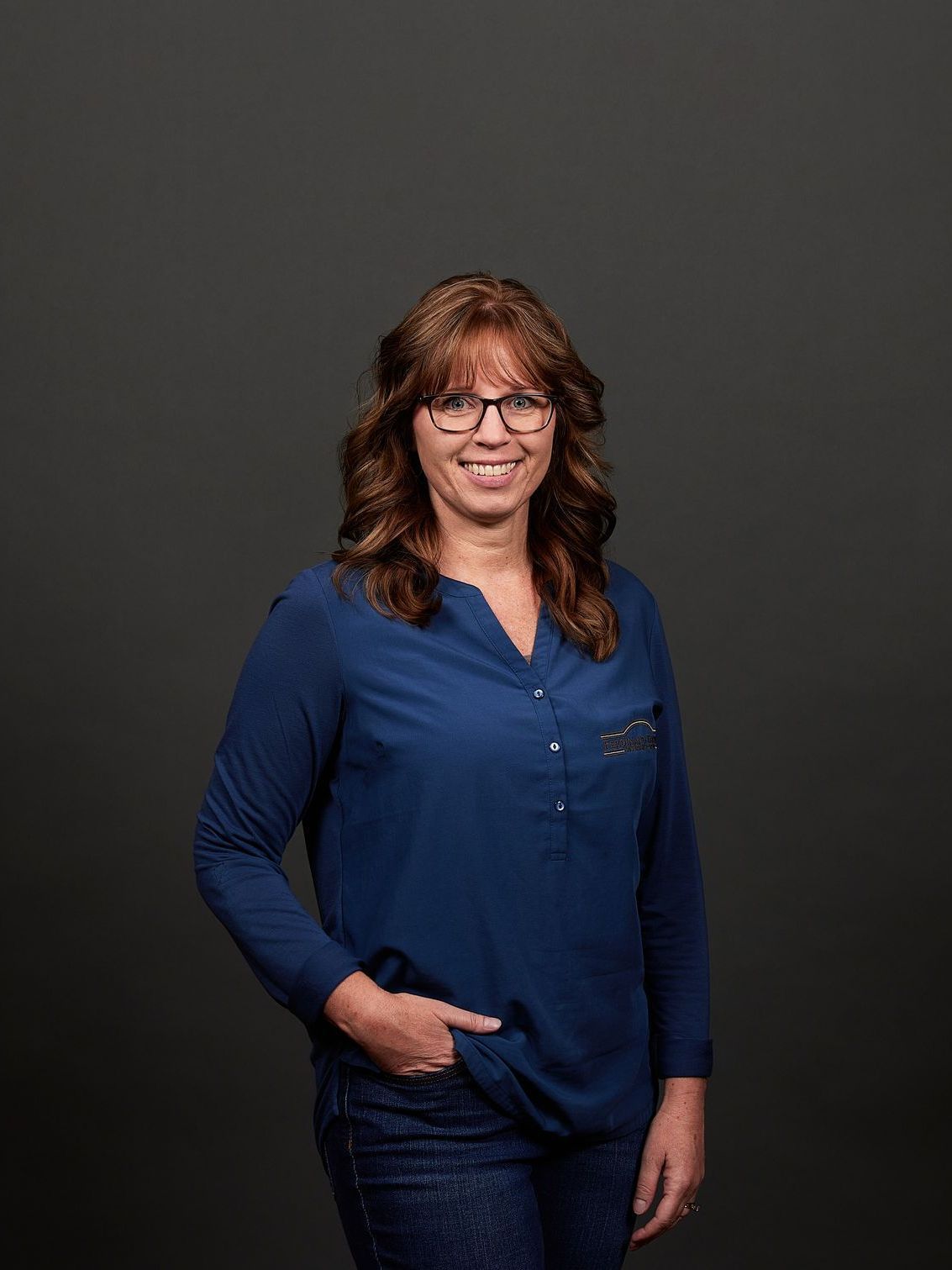 Woman with glasses, dark blue shirt, and jeans, smiling, hand in pocket. Dark gray background.