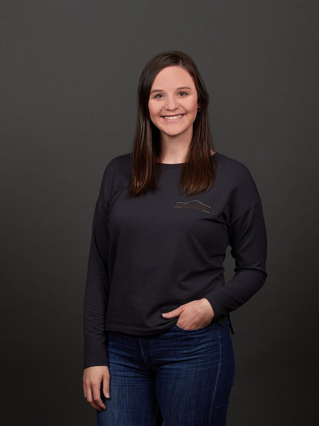 Woman smiling, wearing a black long-sleeved shirt and blue jeans, hand in pocket, gray background.