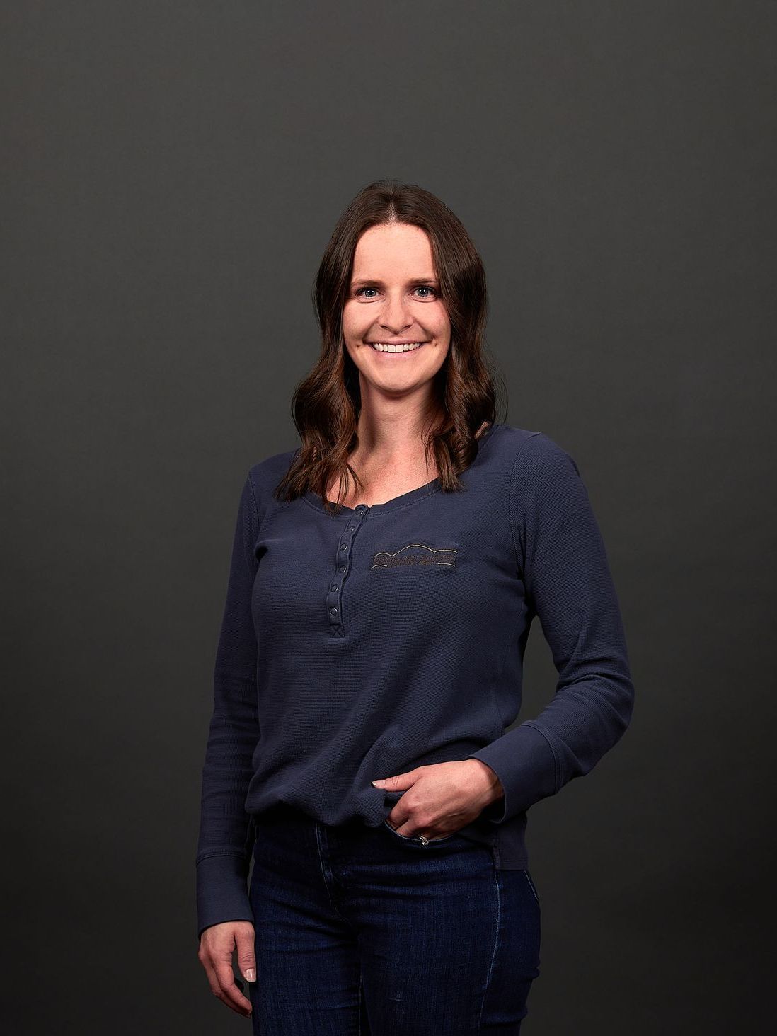 Woman in a blue sweater and jeans smiles, standing against a gray backdrop.
