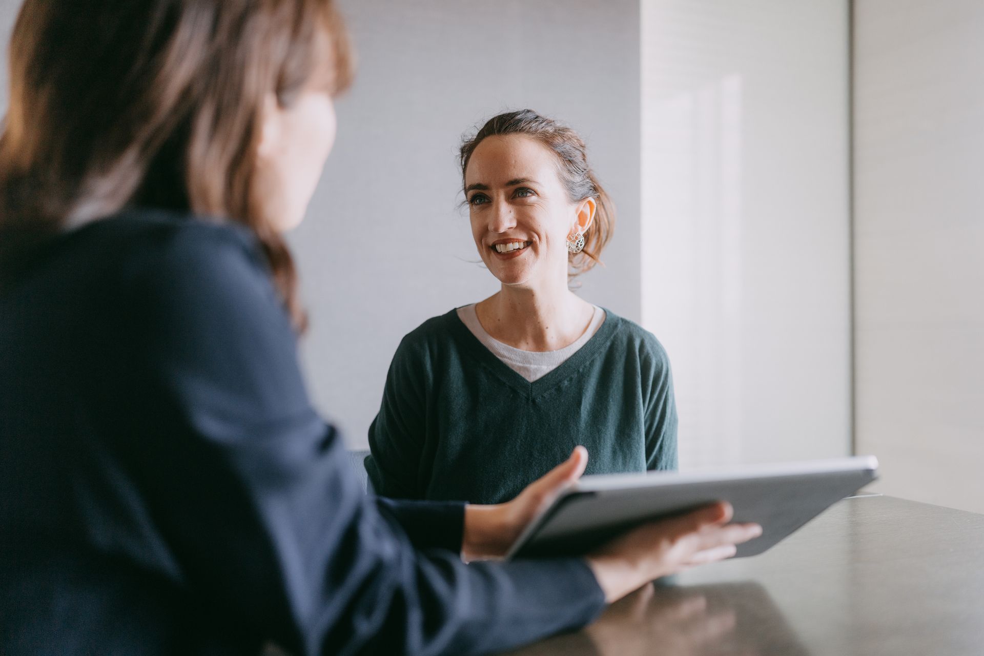 Woman holds tablet, facing another smiling woman indoors.