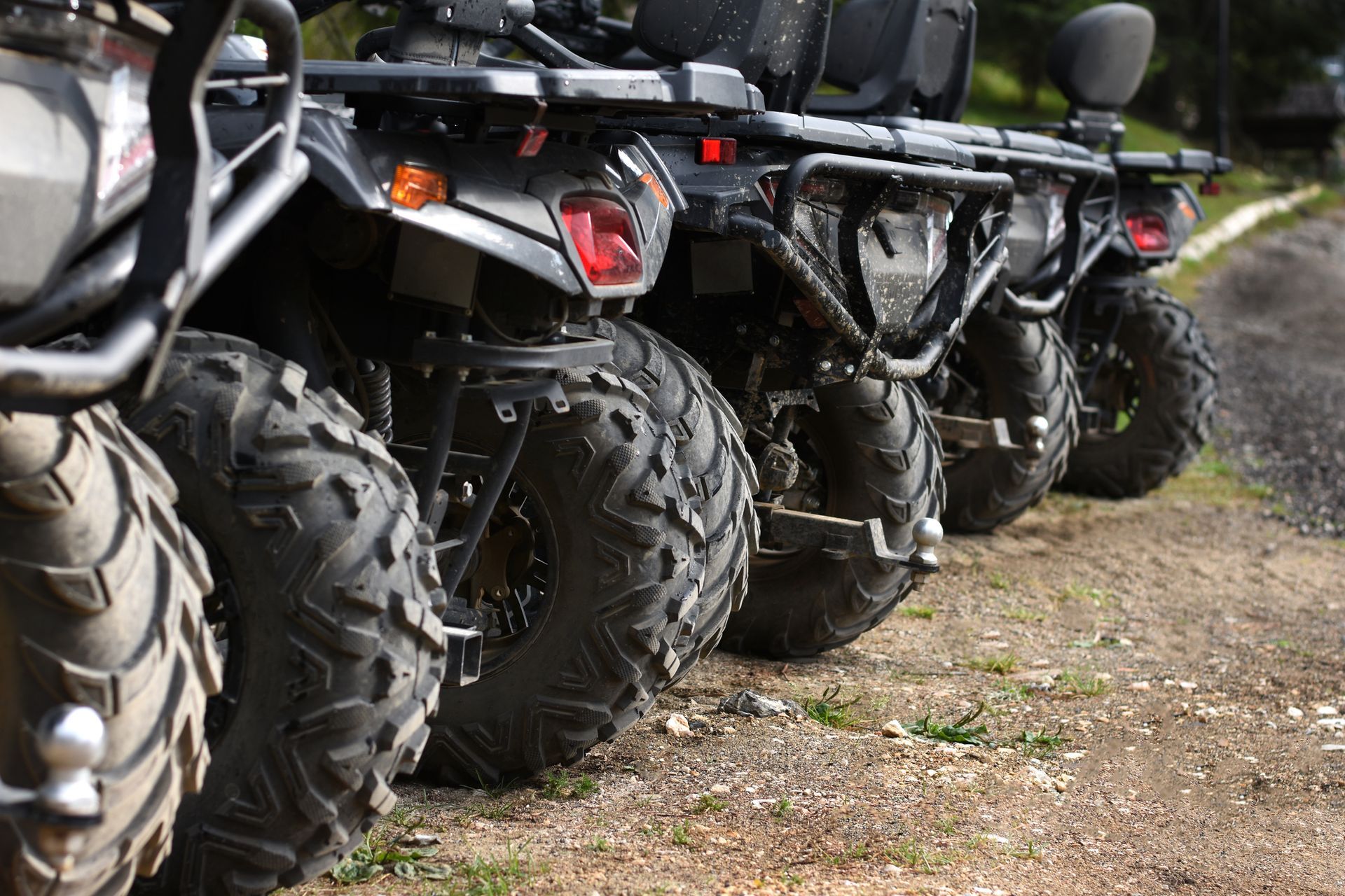 Four black ATVs lined up on a gravel road, showing their tires and rear ends.