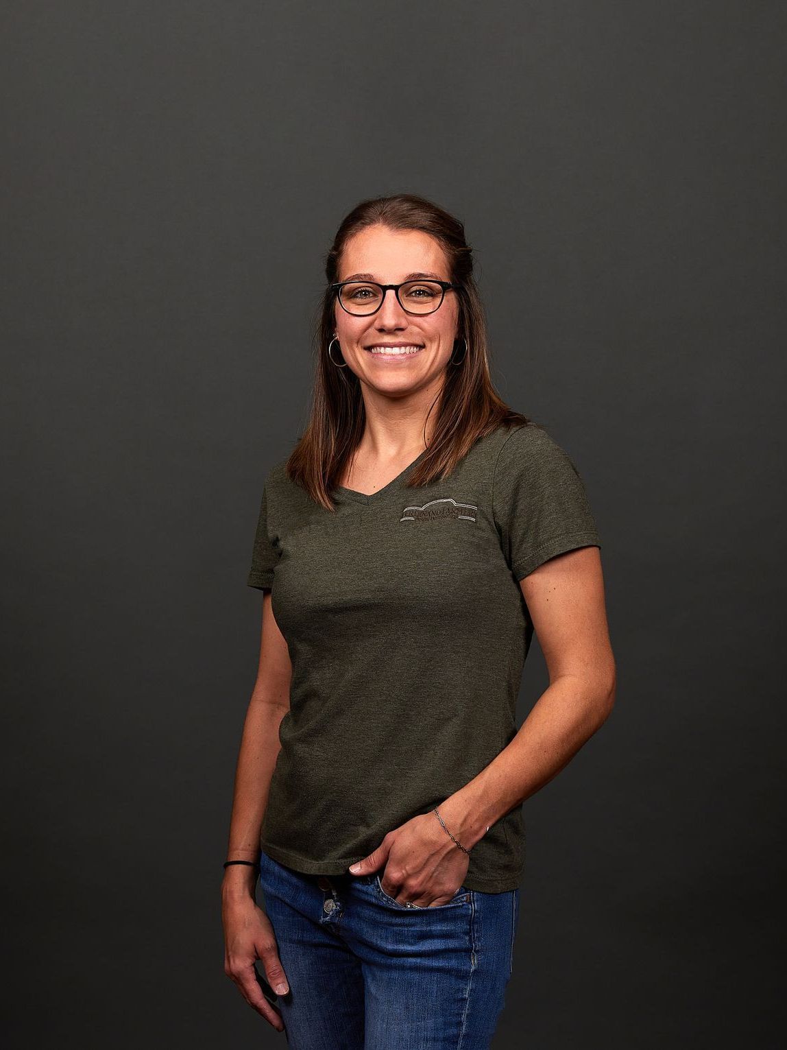 Woman in glasses and green shirt smiles, one hand in pocket, against a gray background.