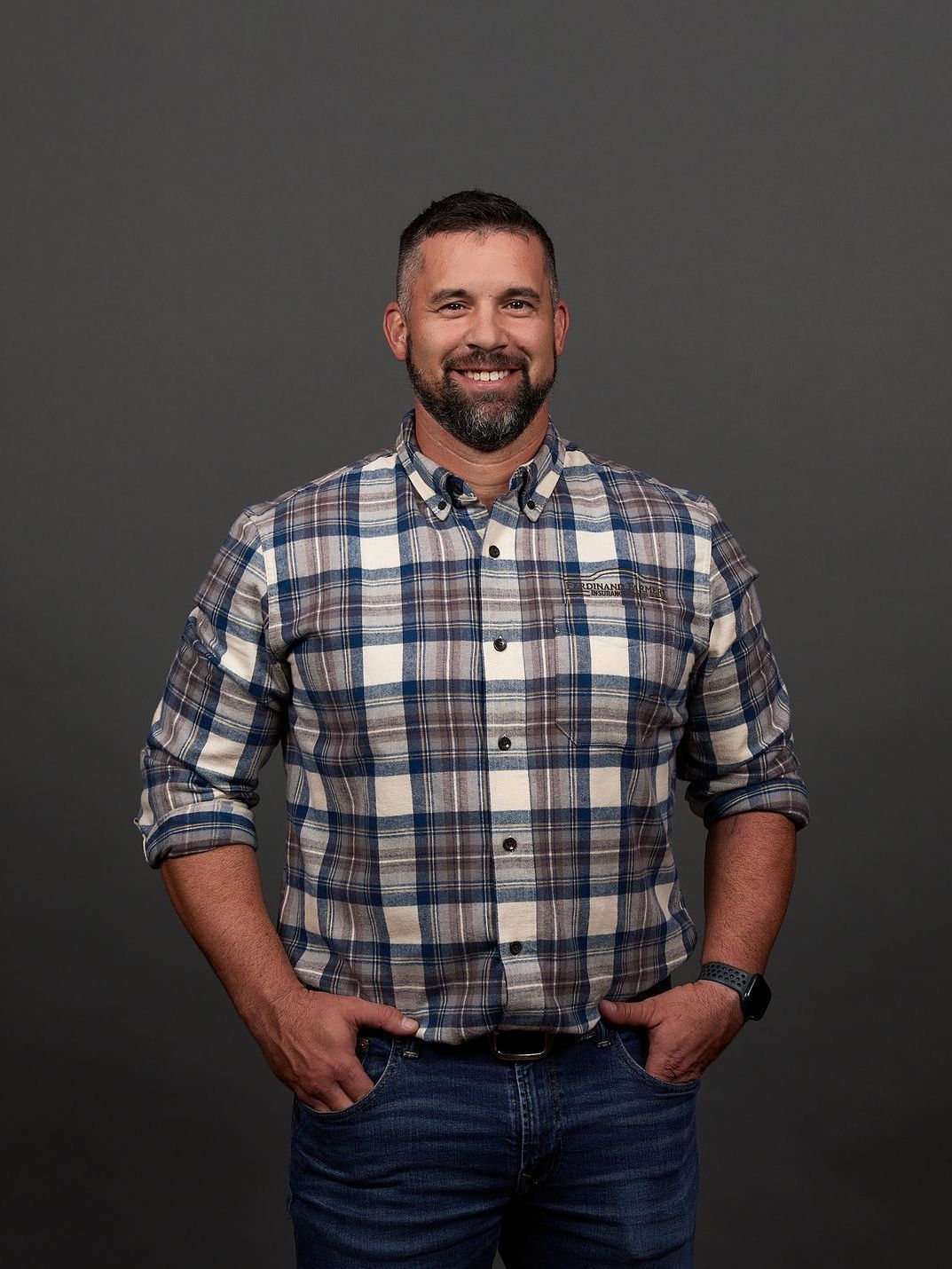 Man with beard wearing plaid shirt and jeans, hands in pockets, smiling.