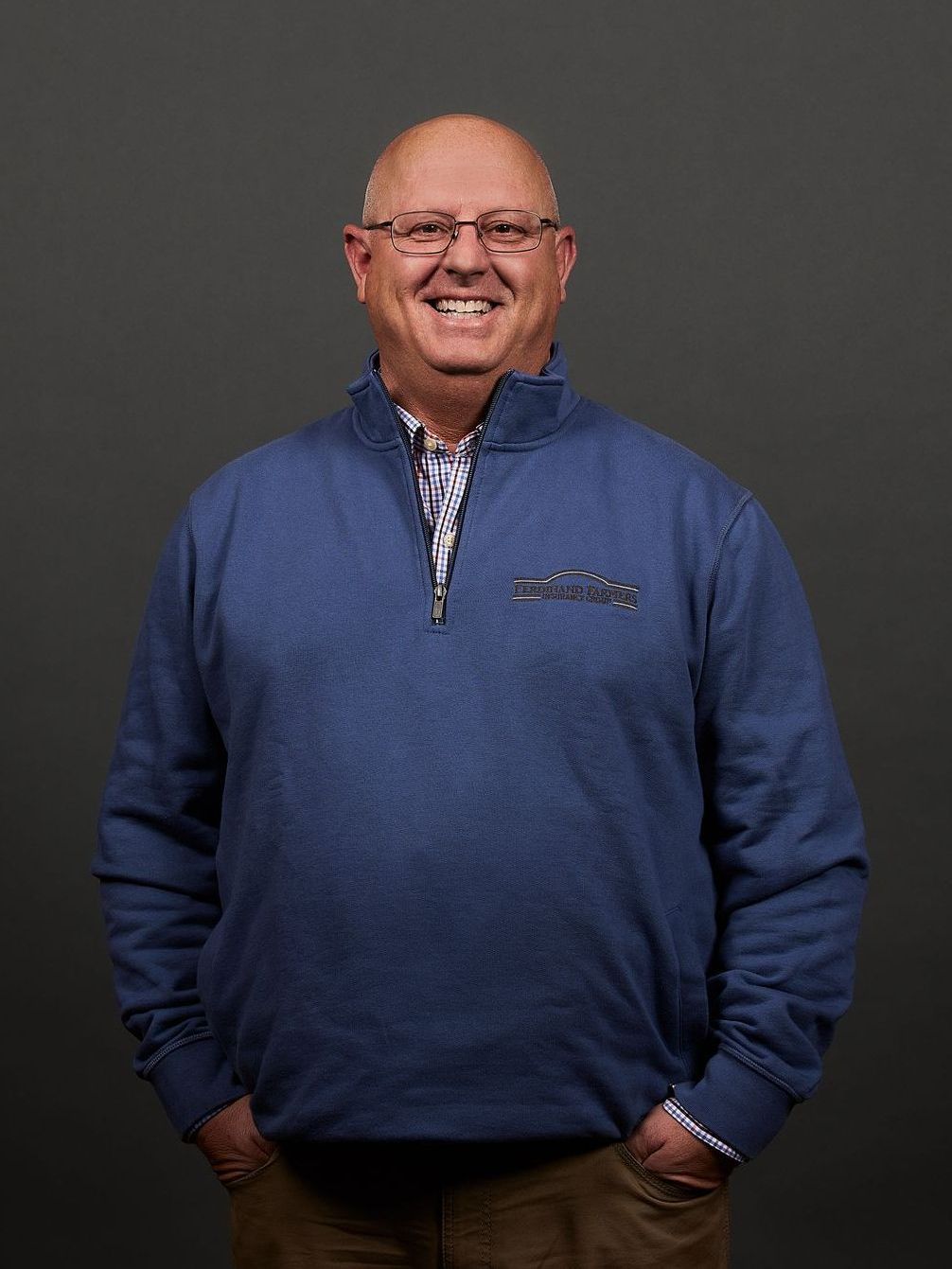 Man in glasses smiles, wearing blue sweater, hands in pockets. Gray background.