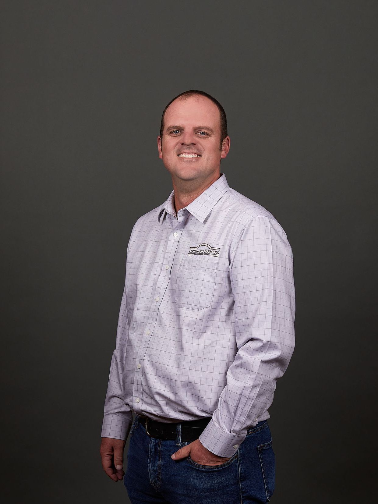Man in light purple button-down shirt and jeans, smiling, hand in pocket, against gray backdrop.