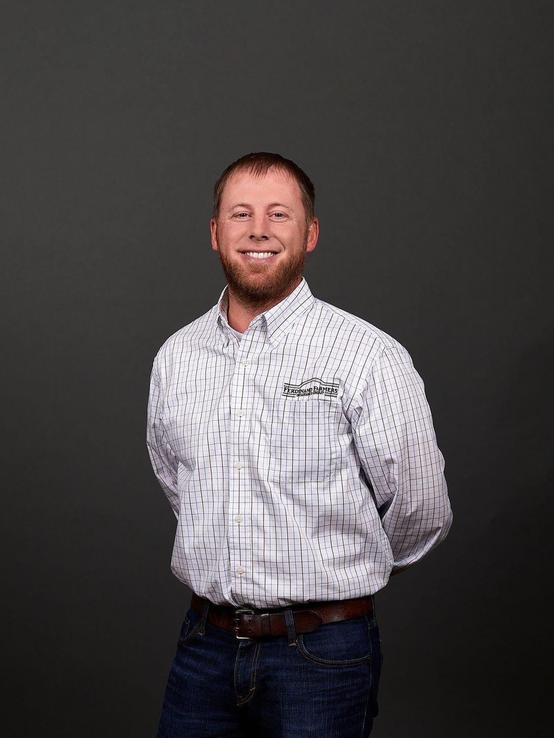 Man with a beard smiling, wearing a patterned shirt and jeans, hands clasped behind his back.