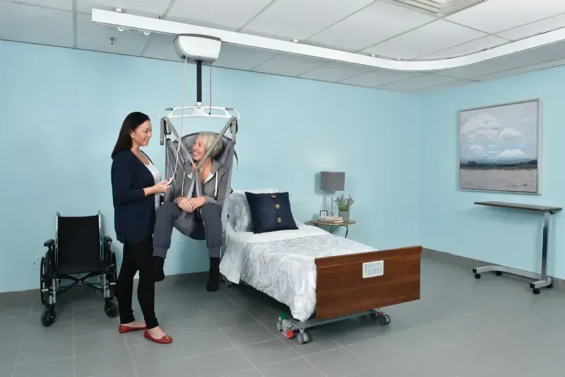 A woman is helping an elderly woman in a hospital bed using a patient lift installed by Altamira Ltd.