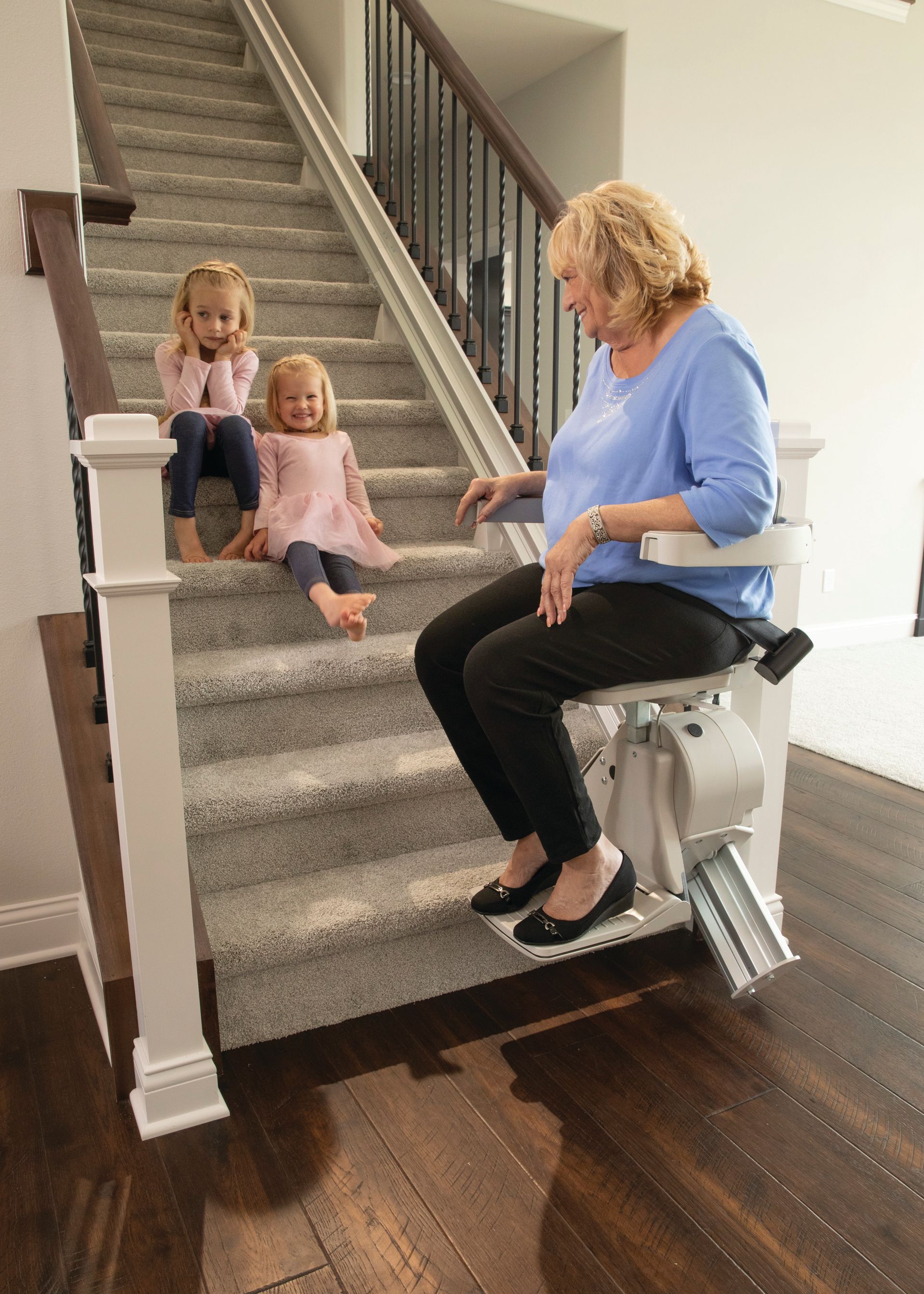 A woman is sitting on a stair lift with two little girls sitting on the stairs.