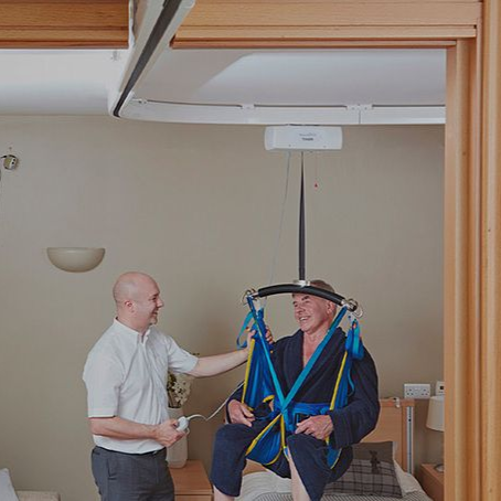 Man in harness being assisted by another in a room, using a ceiling lift.