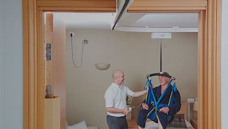 Man in harness being assisted by another in a room, using a ceiling lift.