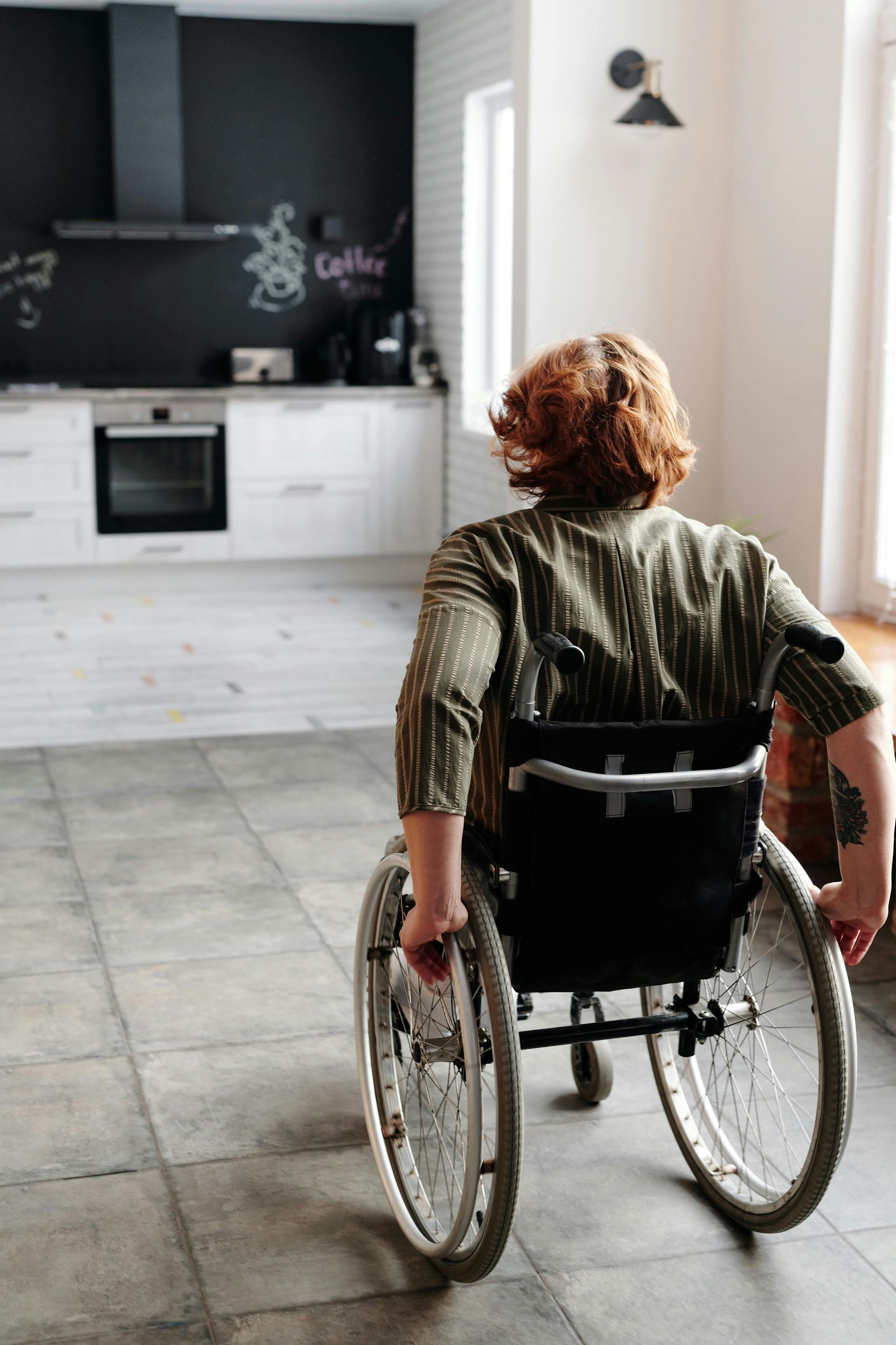 A woman in a wheelchair is sitting in a kitchen looking out a window.