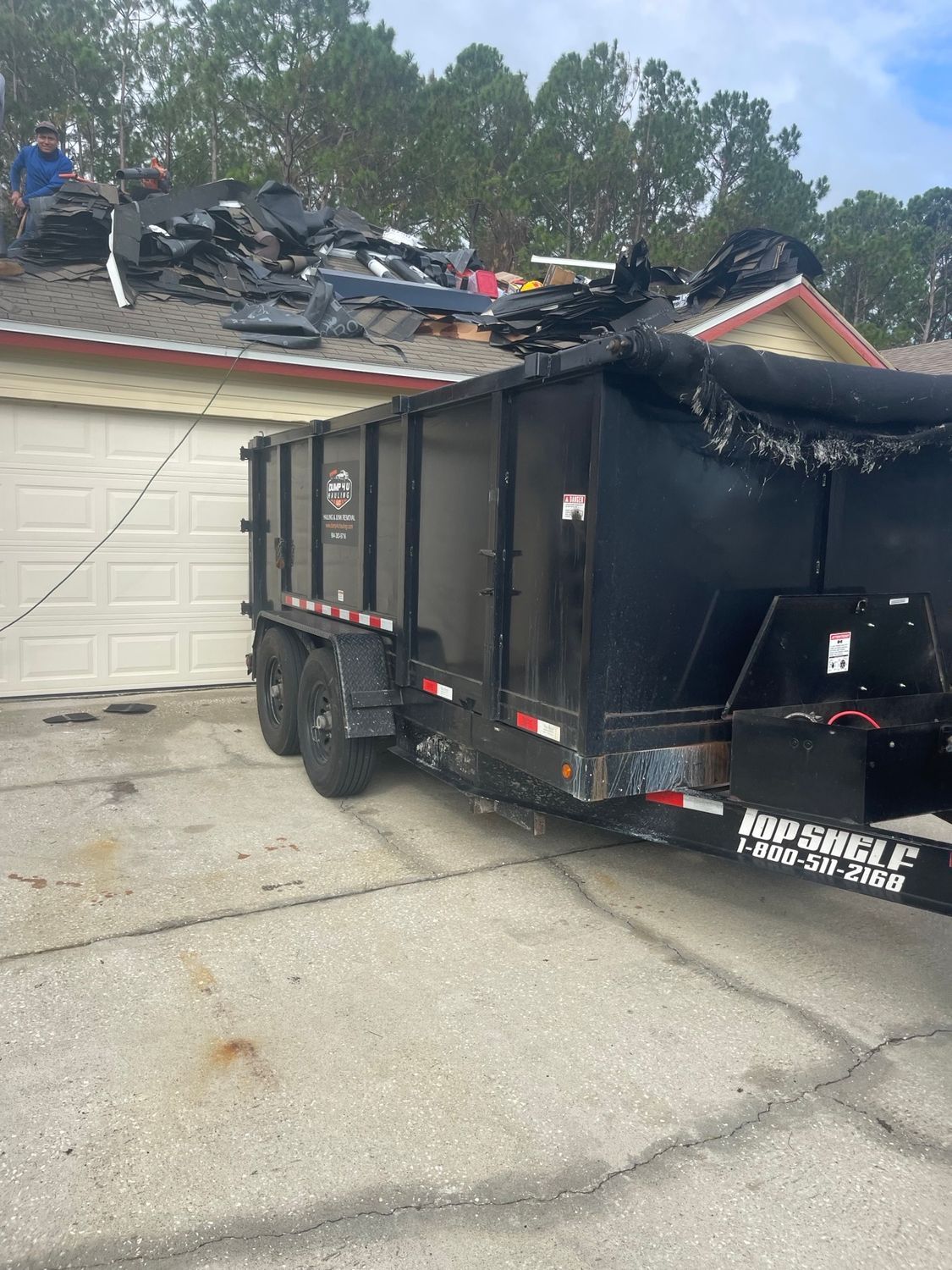Yellow dumpster on a street next to a brick building and a gray truck on a sunny day.