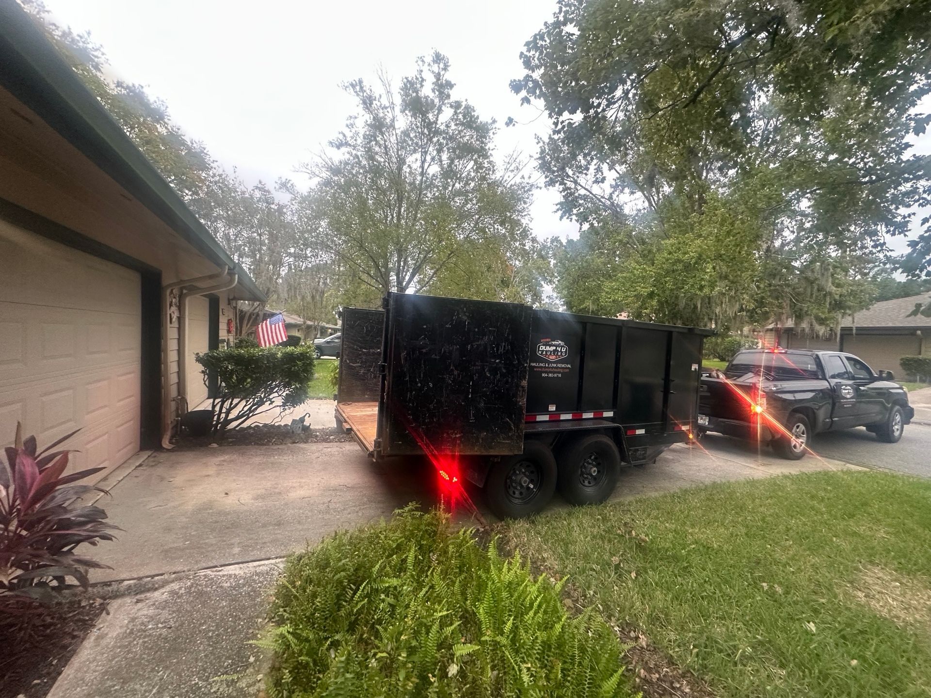Black trailer on a street next to a blue house with a white fence and a portable toilet.