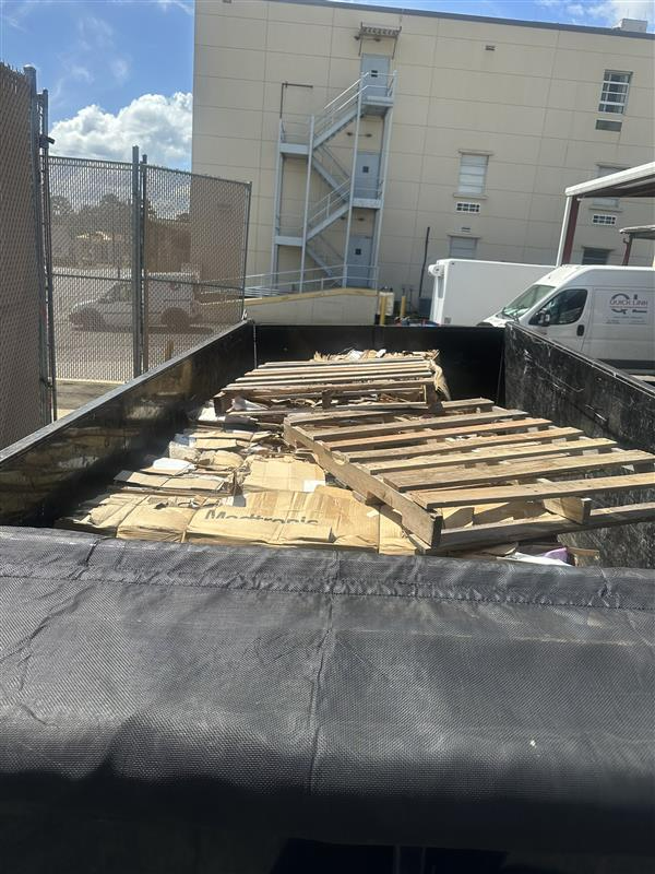 Blue dumpster overflowing with trash, next to a concrete wall and a curved sidewalk.