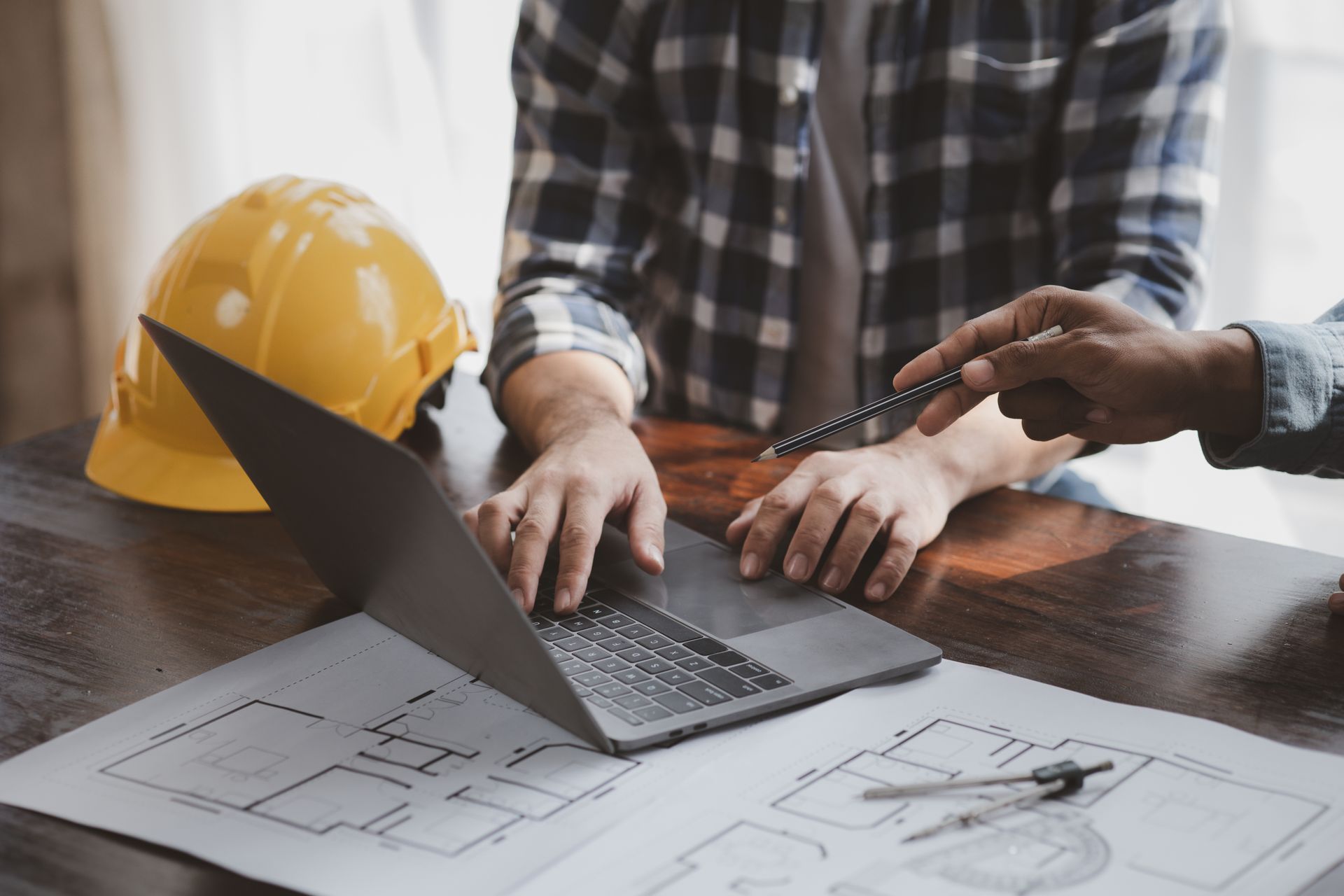 Two home designers are reviewing a house plan on a blueprint and laptop. A hard hat is to the side.
