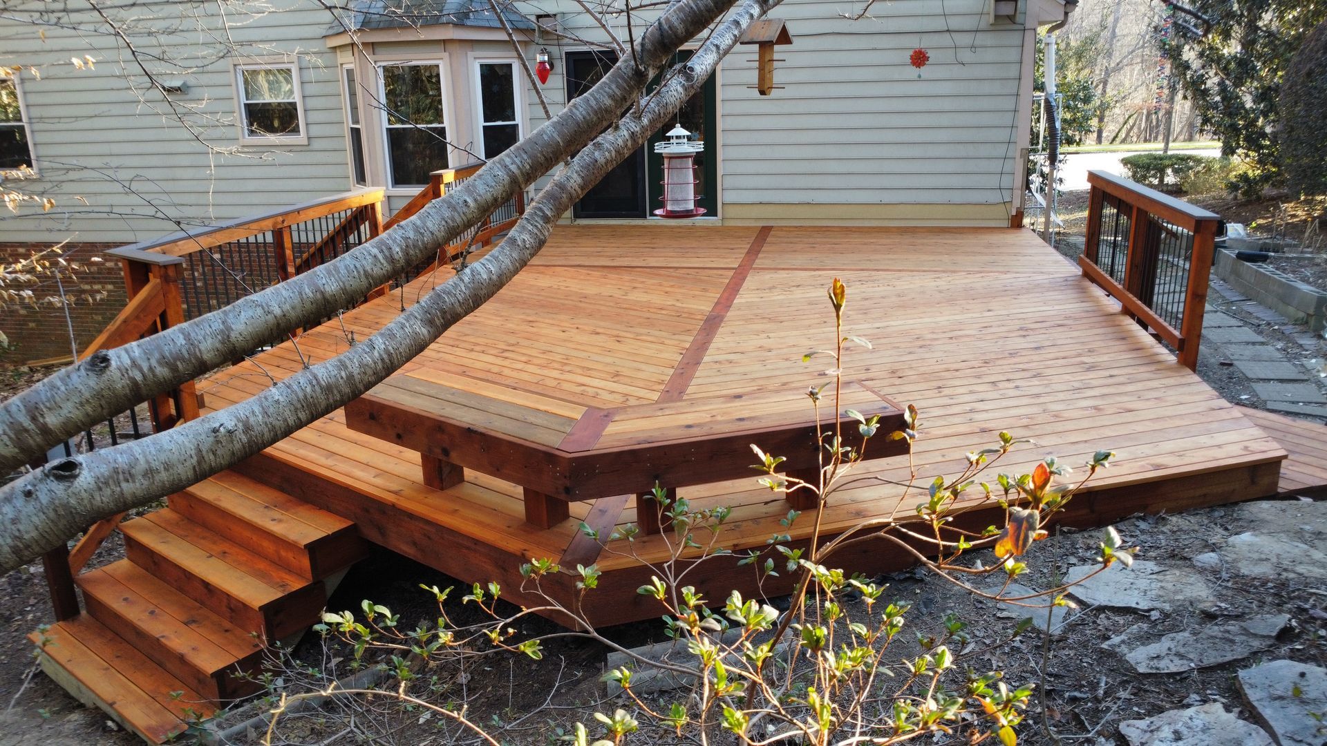 Wooden deck with built-in bench, steps, and railing. Tree branches in front, next to house.