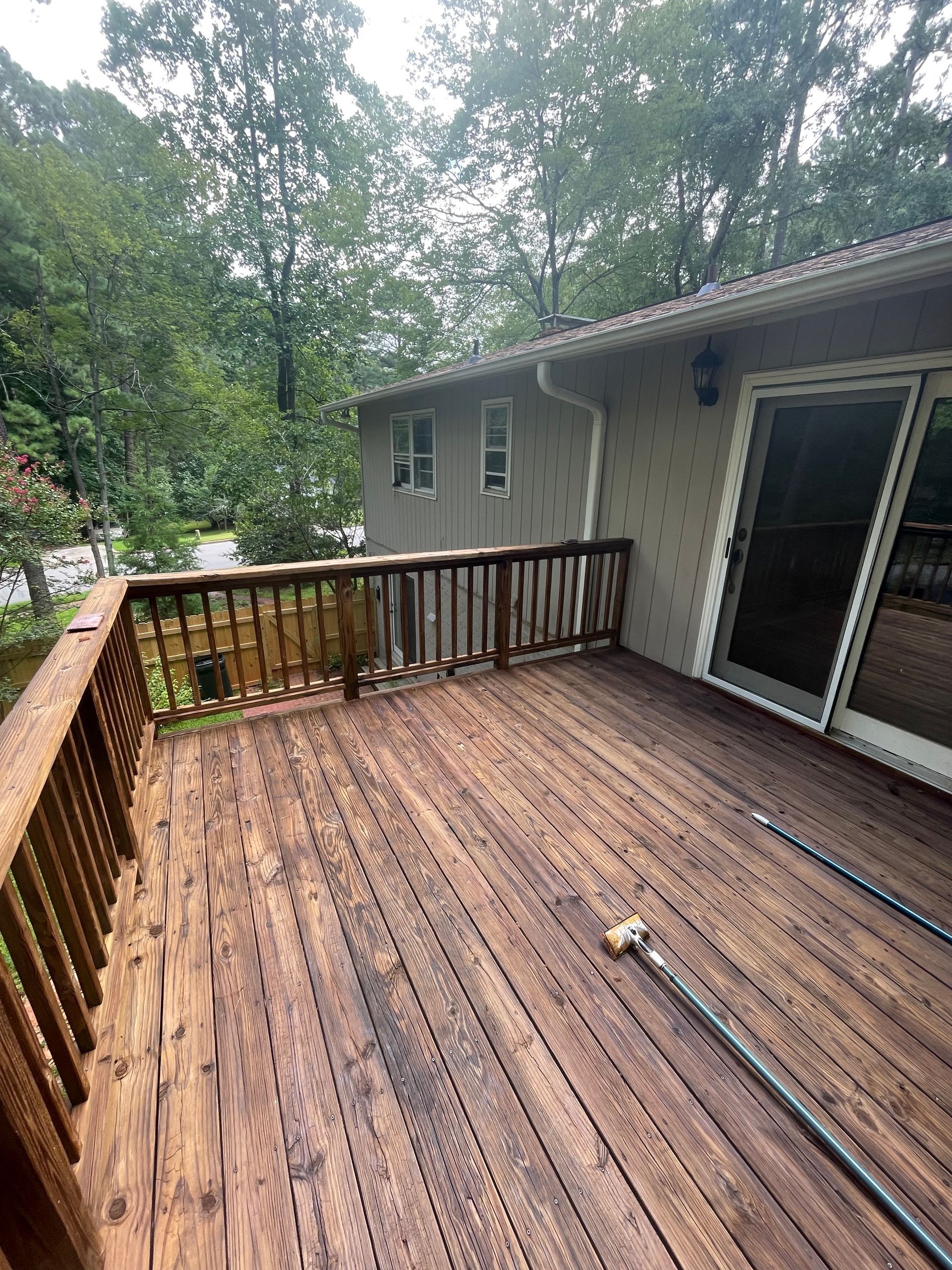 Wooden deck with railing, connected to a beige house, hose visible. Green trees in background.