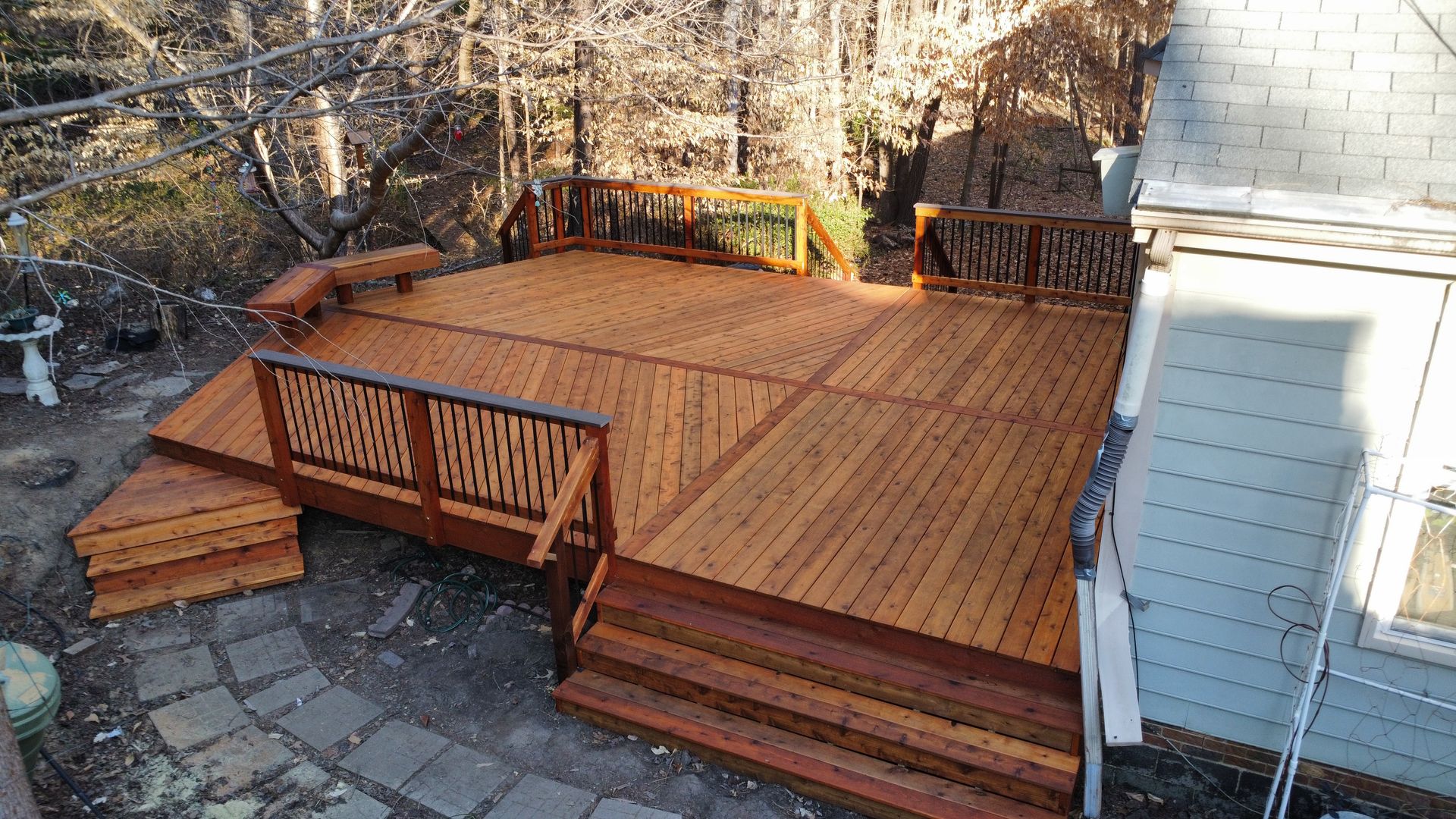 Wooden deck with stairs, benches, and intricate railings, beside a house with gray siding, outdoors.