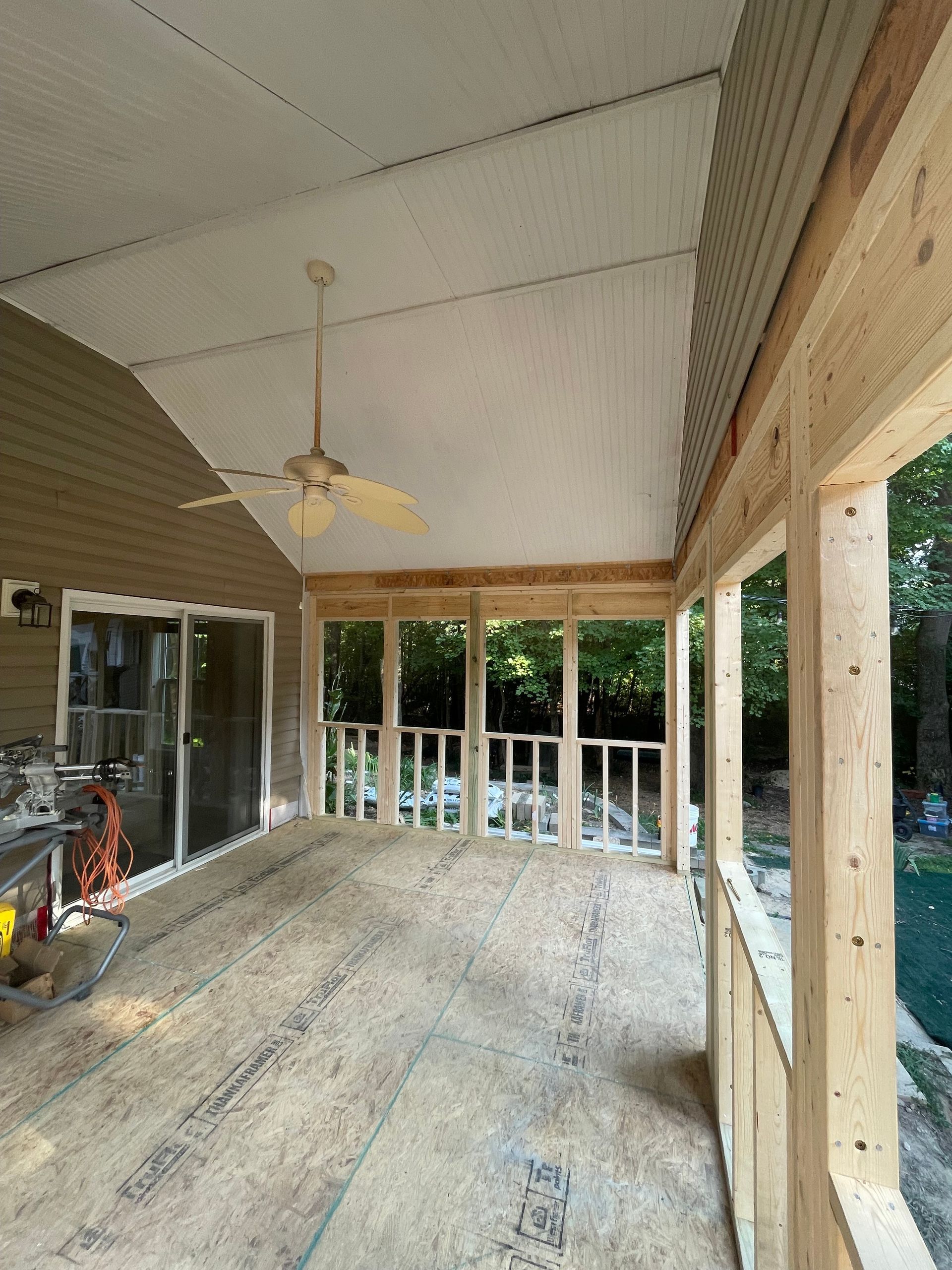 Unfinished screened porch with wood framing, ceiling fan, sliding door, and lumber on the floor.