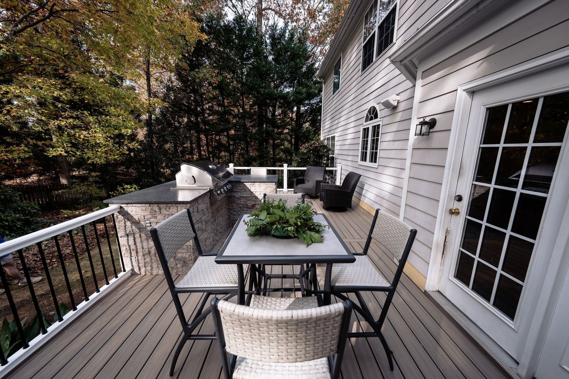 Deck with outdoor dining set and grill, next to a house with trees in the background.
