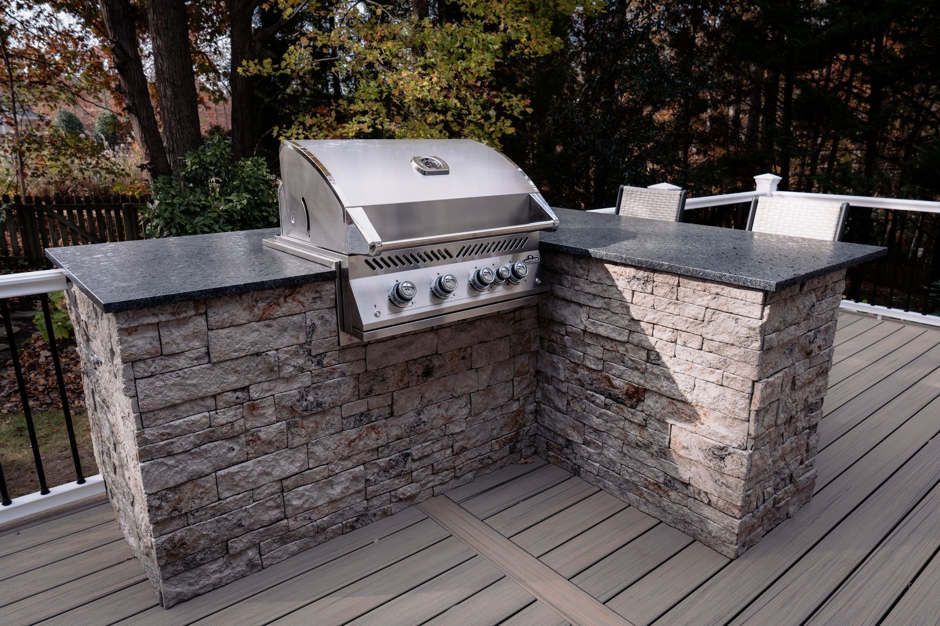 Outdoor kitchen with a stainless steel grill, stone facade, and dark countertop on a wooden deck.