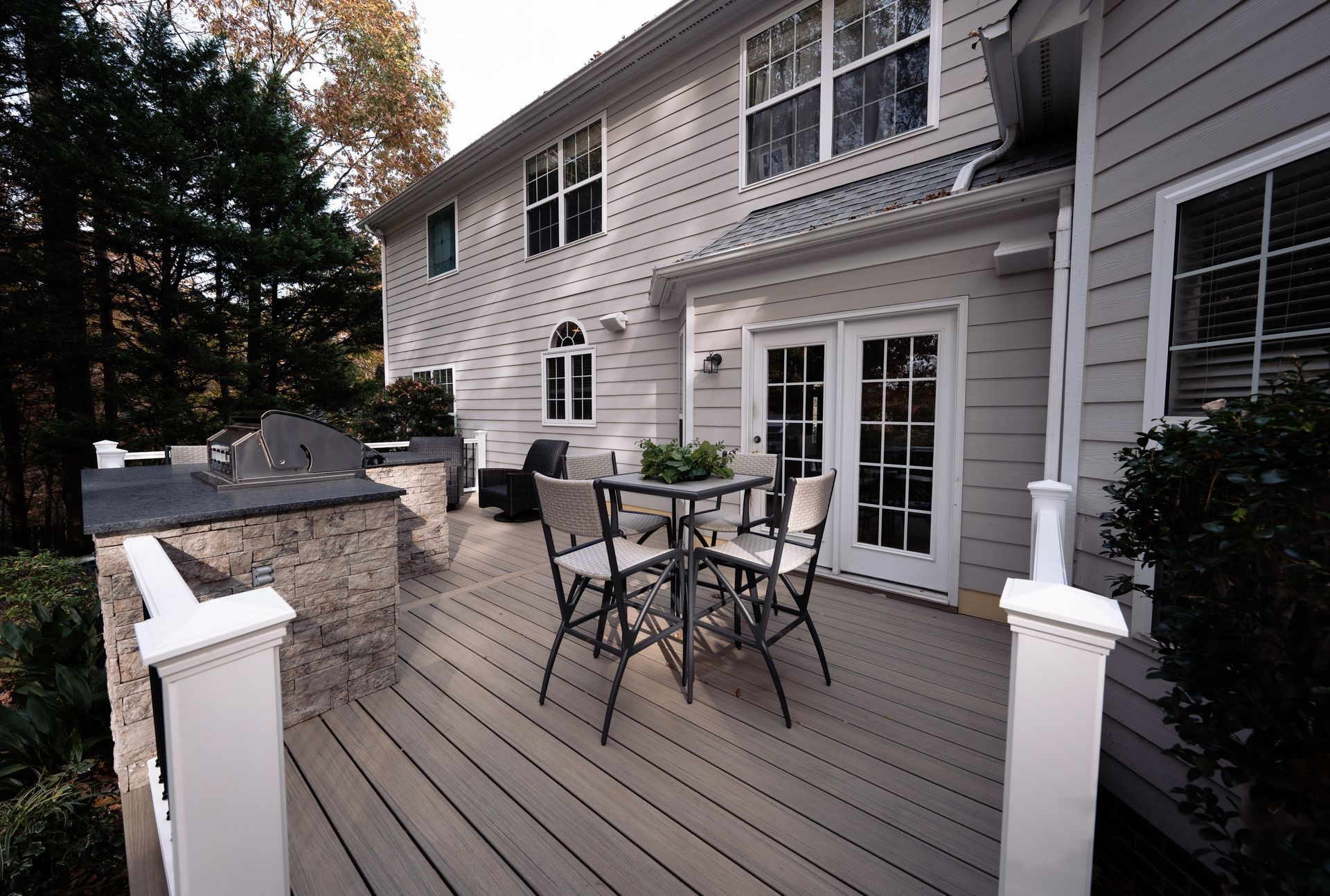 Deck with table, chairs, and grill, next to a two-story house with French doors.
