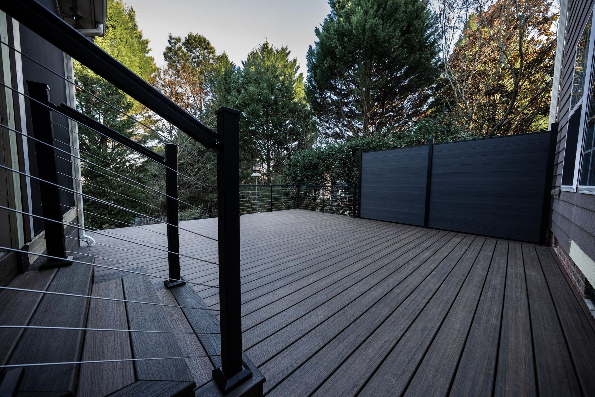 A dark wood deck with black railings and cable balusters, featuring a private solid wall panel overlooking green trees.