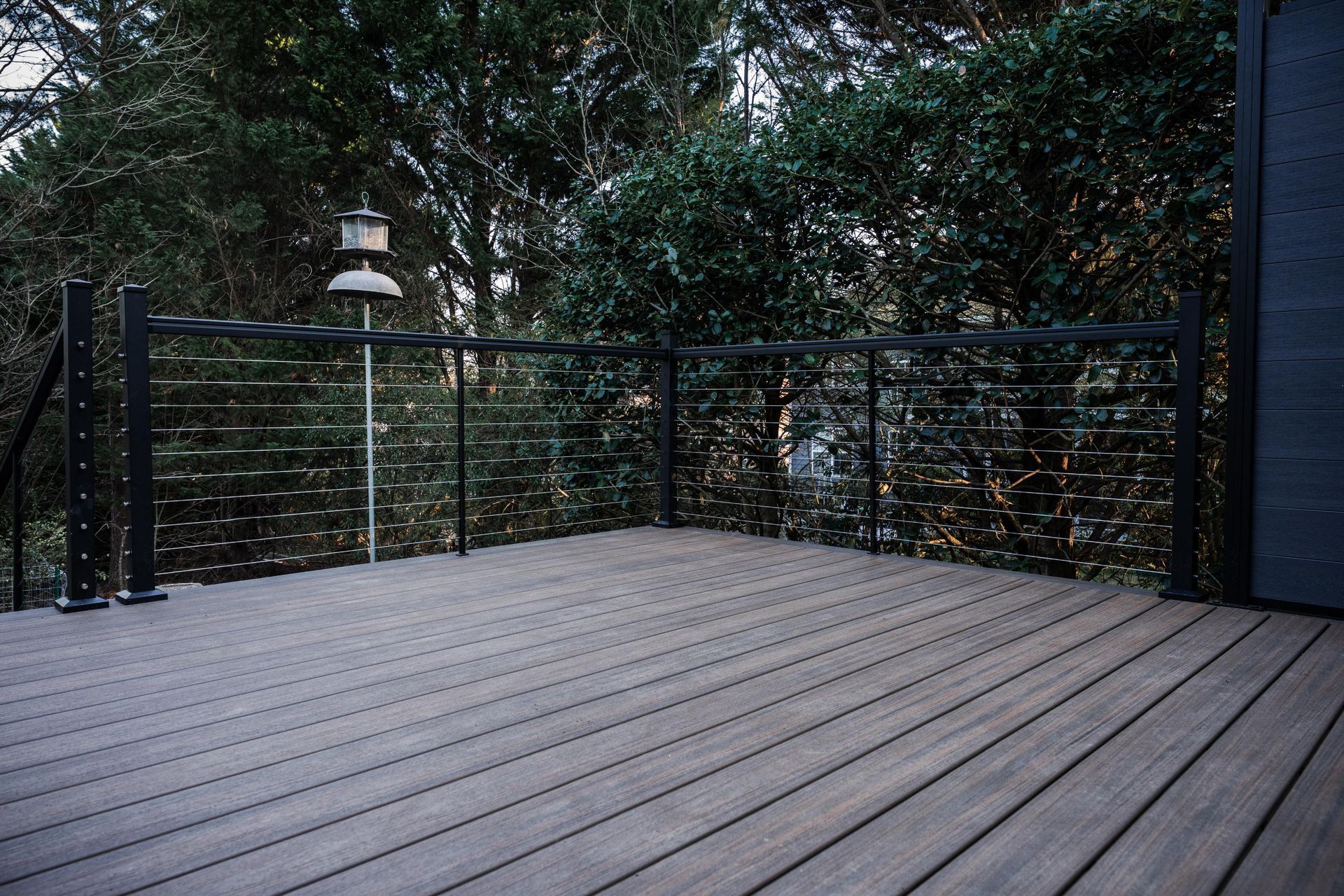 A wooden deck with a black metal cable railing overlooks dense trees in an outdoor setting.