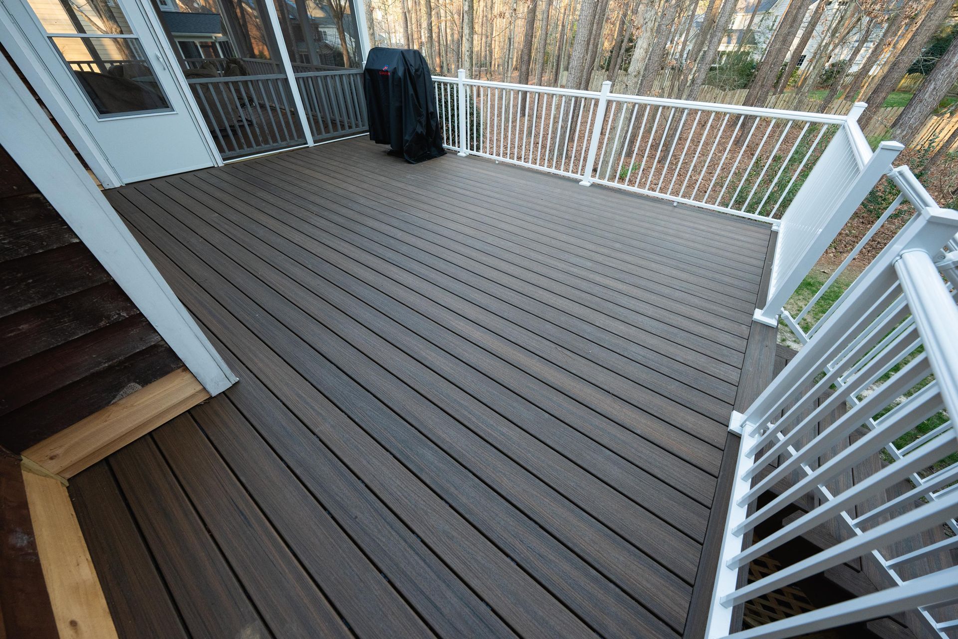A dark brown composite deck with white railings, featuring a covered grill in the corner against a wooded backdrop.