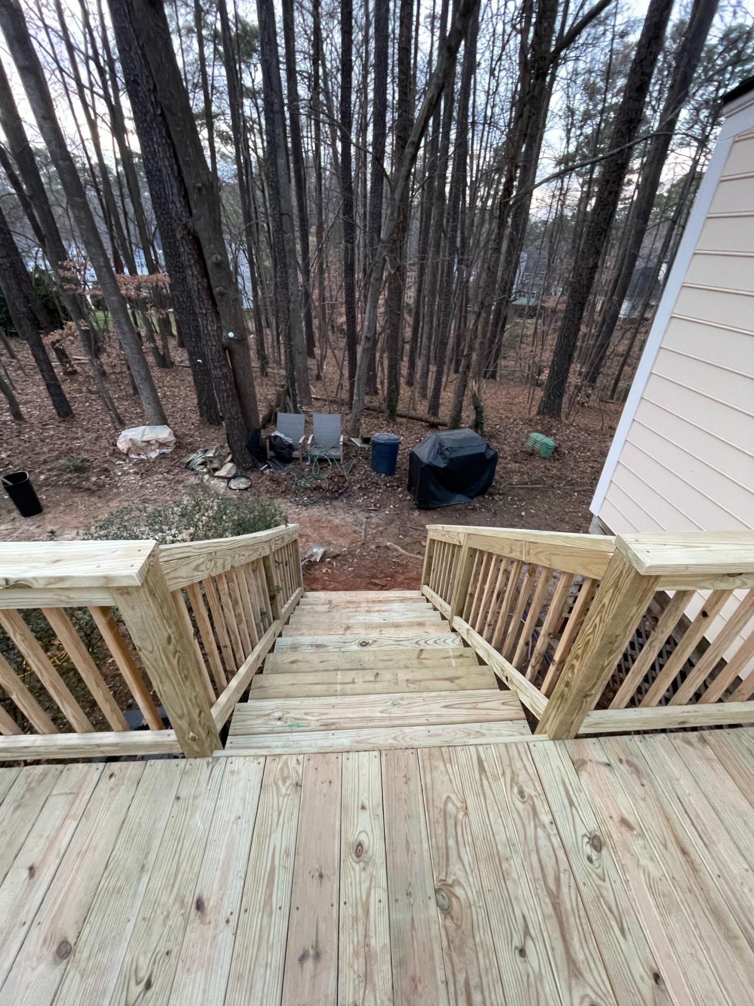 A wooden deck staircase leads down to a wooded backyard featuring chairs, a grill, and tall trees.