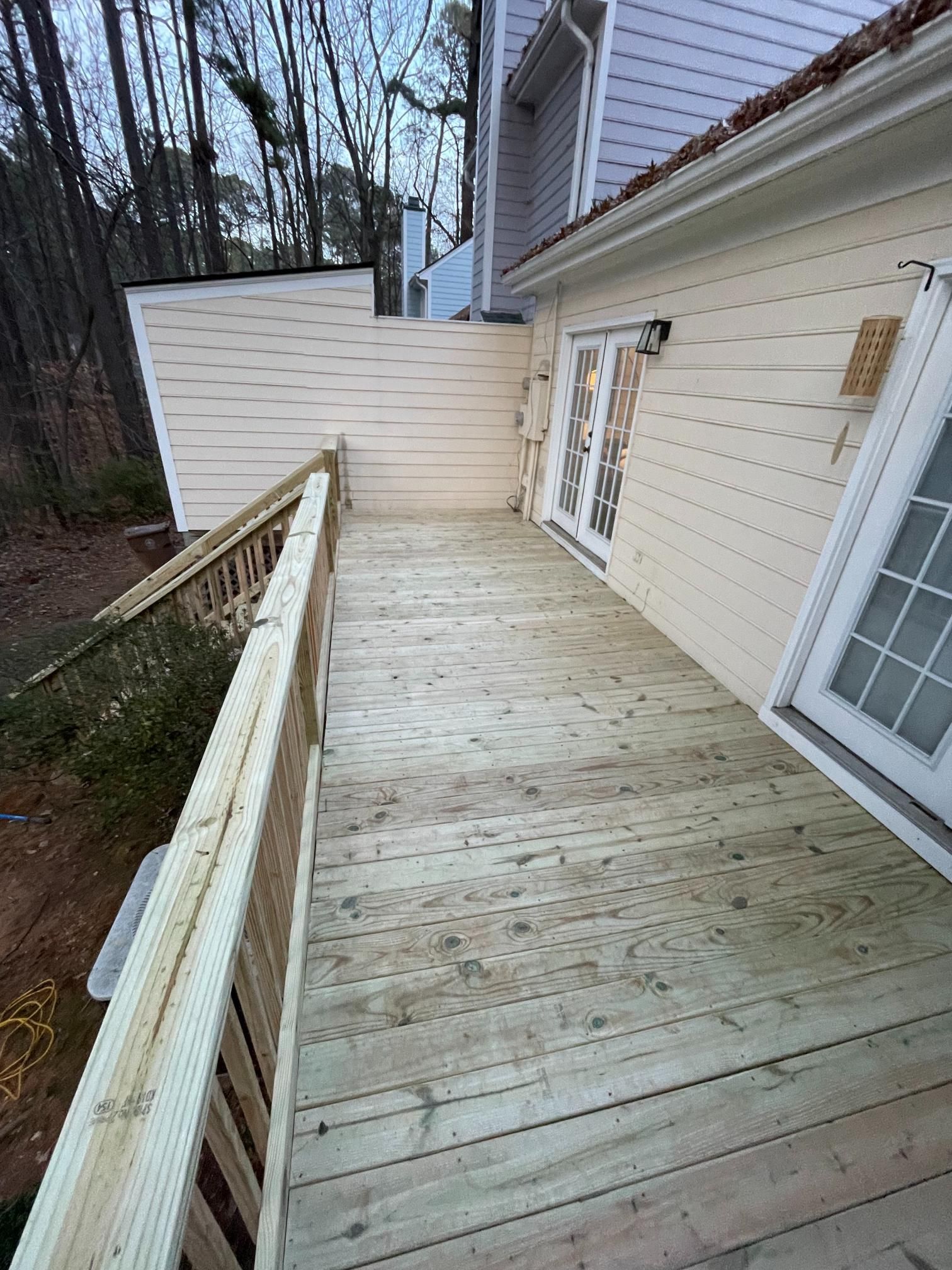 A newly constructed wooden deck featuring a railing alongside the side of a light-colored house with glass doors.