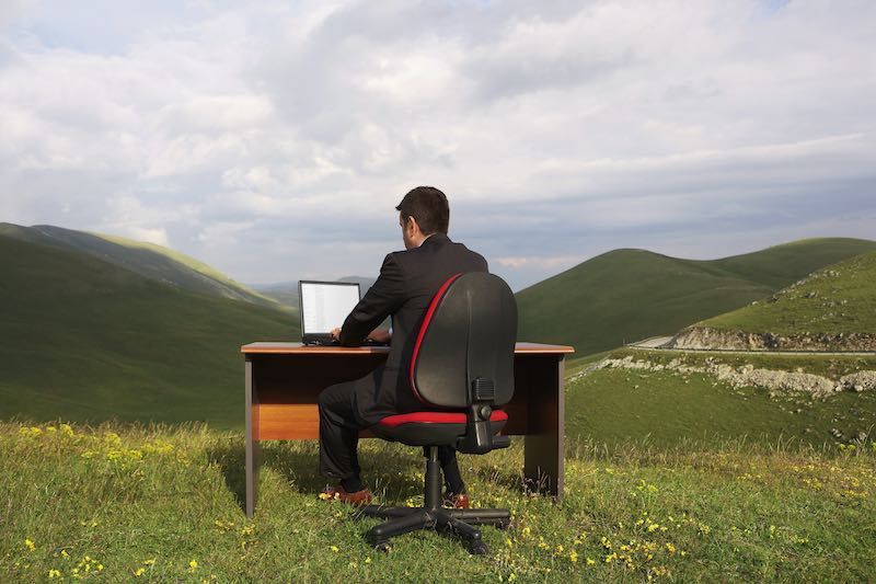 Man in suit at desk, using laptop in a grassy field with rolling green hills and cloudy sky in the background.