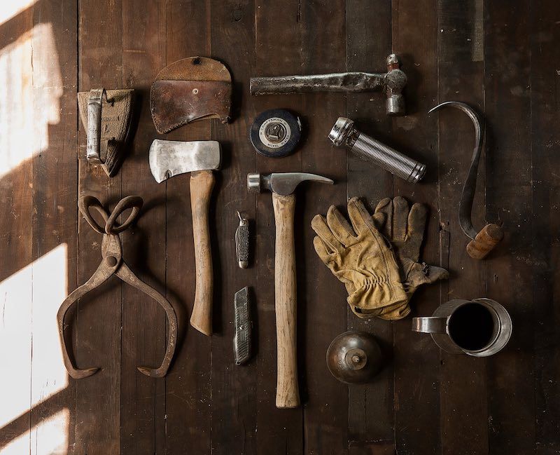 Tools arranged on a dark wood surface: axe, hammers, gloves, tongs, cup, and other hardware.