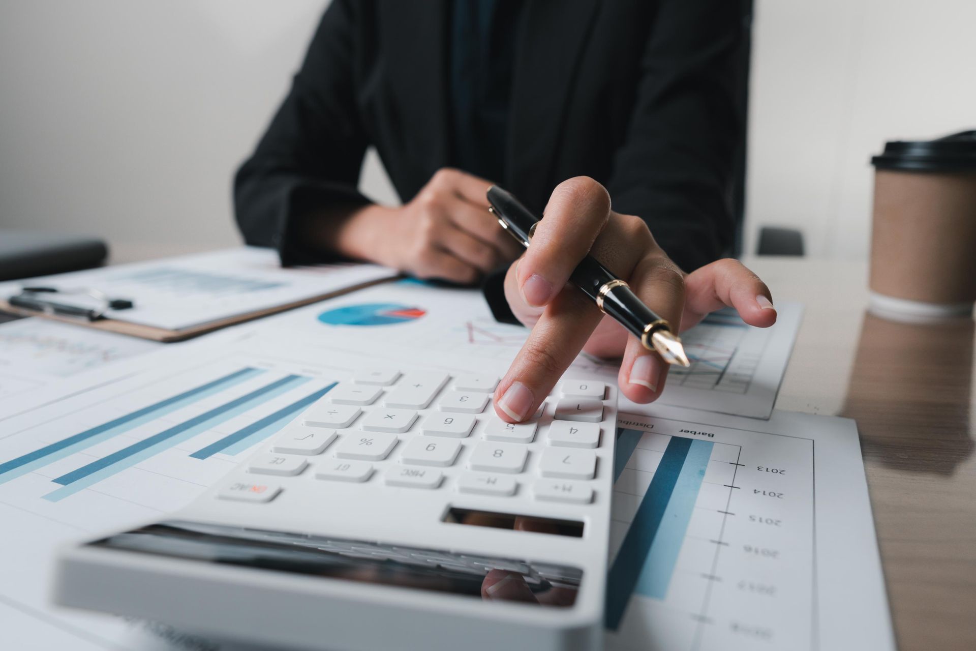 Person in black blazer using a calculator, looking at financial charts with a pen nearby.