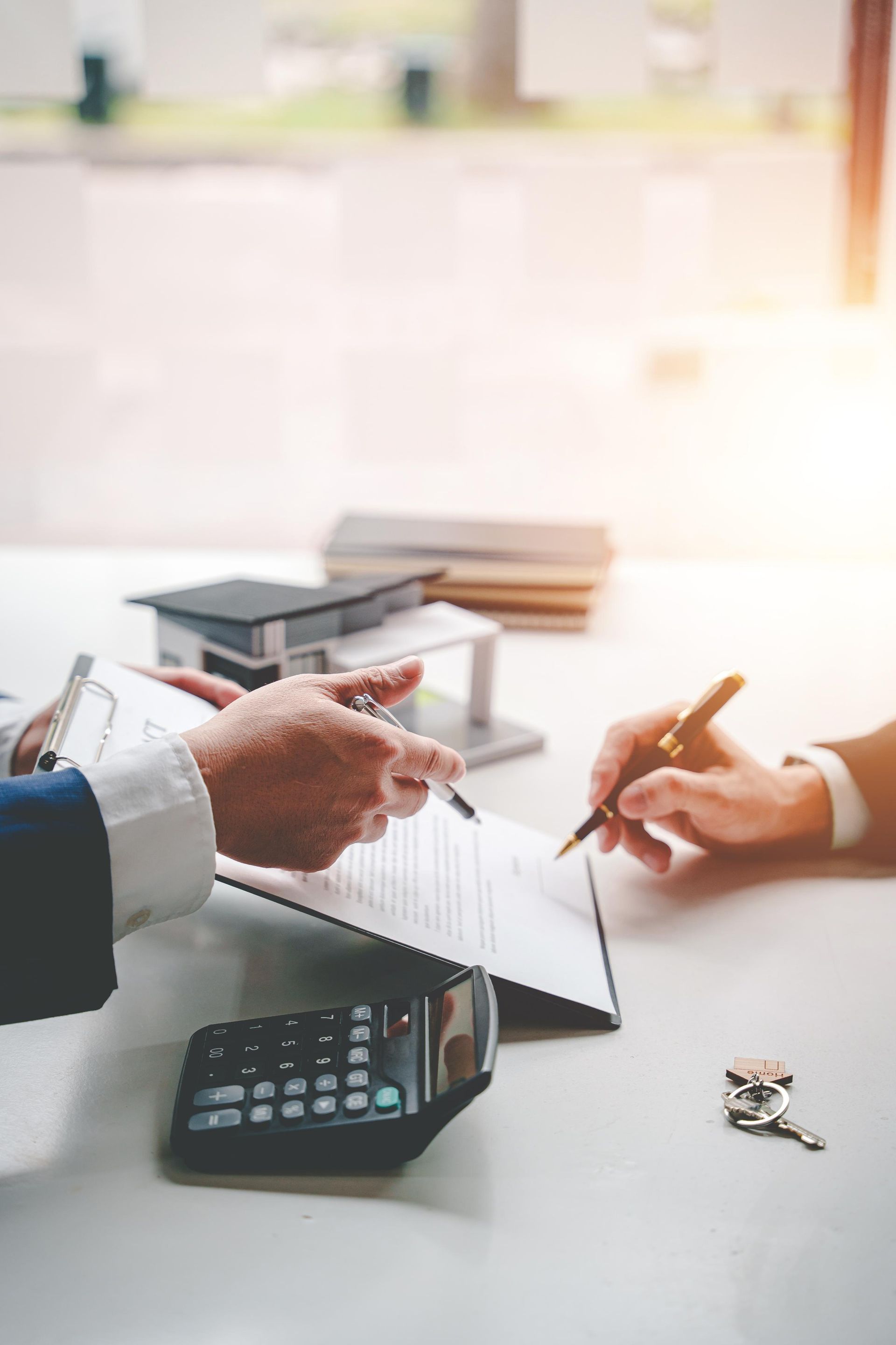 Hands signing a document, likely a real estate contract, with a house model and keys on a table.